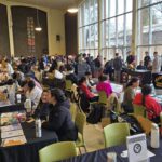 The image shows a room full of Chabot College students gathered around rows of tables draped in black tablecloths. Some students sit behind the tables and are giving information about various school clubs to the students who are standing in front of them.