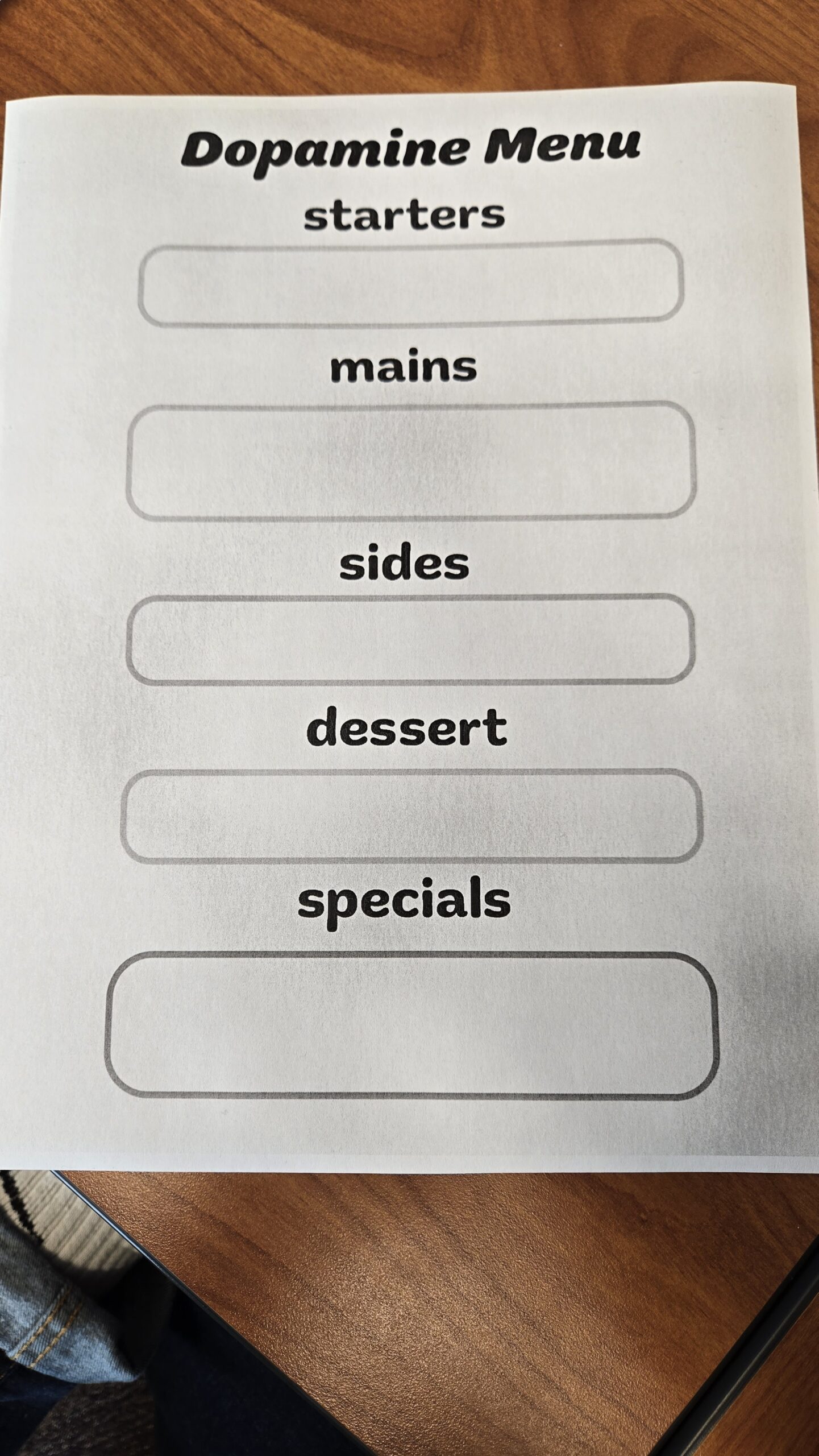 A printed worksheet titled “Dopamine Menu” on a wooden surface. The worksheet is structured like a restaurant menu with sections labeled “starters,” “mains,” “sides,” “dessert,” and “specials,” each containing an empty box for writing. The sheet is designed to help individuals categorize activities or habits that boost dopamine levels in a fun and engaging way.