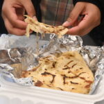 A pupusa being pulled apart with cheese falling out, aluminum in foil in a tray