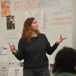 a woman lectures in front of an audience with a projector screen and whiteboard in background