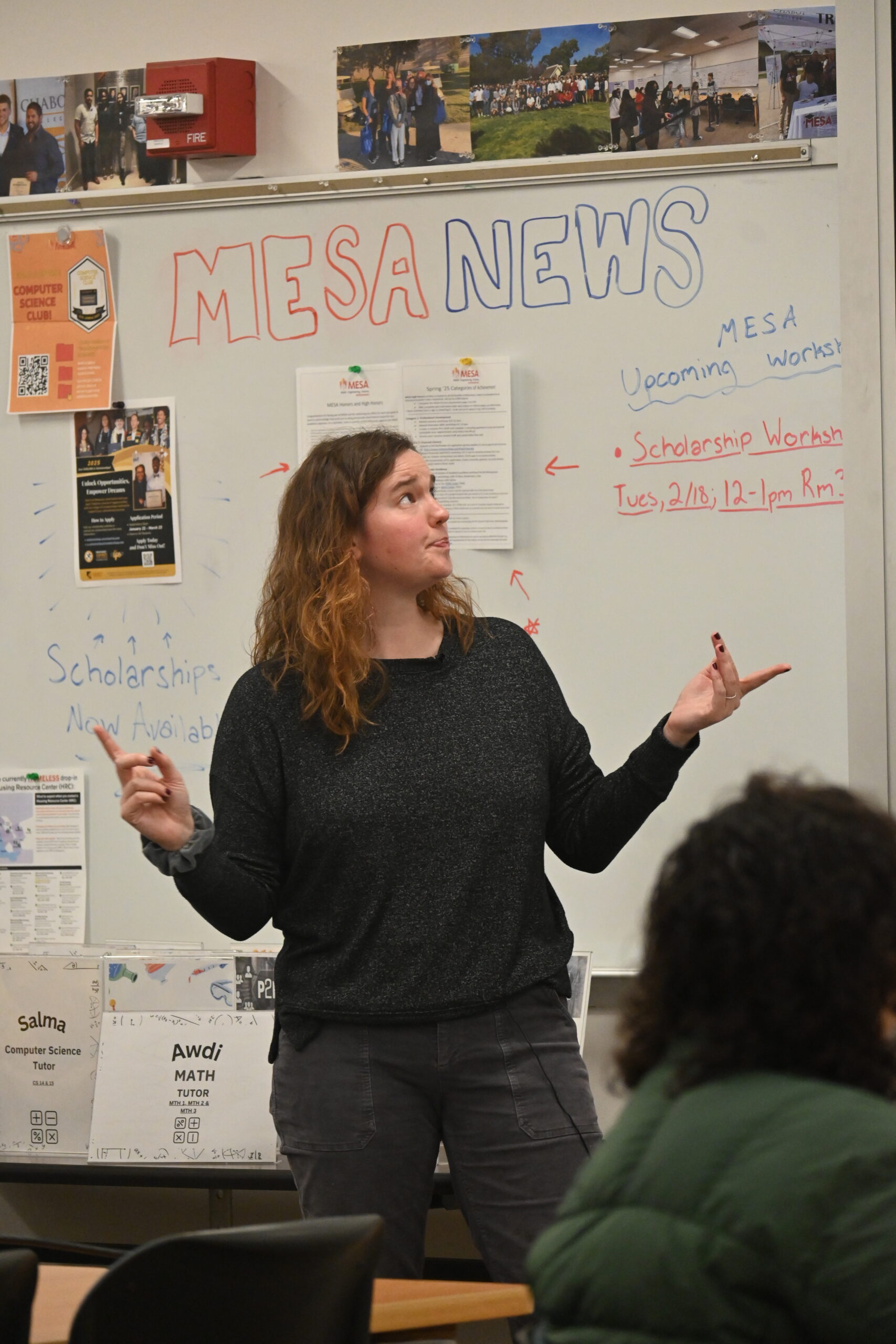 a woman lectures in front of an audience with a projector screen and whiteboard in background