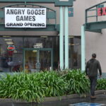 A man is walking up to a storefront in the rain. The storefront is green with large windows, and has a large white sign over the door with black text that reads "Angry Goose Games" in large font. Below that it says "Grand Opening" in a smaller font.
