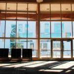 The photo shows the inside of a library with large glass windows, letting in sunlight and shadows. Another modern library building is visible outside, along with an outdoor seating area.