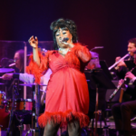 A woman wearing a vibrant red dress with feathered sleeves and hemline sings passionately into a microphone on stage. She has styled dark hair and is accessorized with earrings and a sparkling necklace. Behind her, a live band of musicians in black suits plays instruments including drums, trumpet, clarinet, and saxophone. The stage is warmly lit, creating a lively and elegant concert atmosphere.