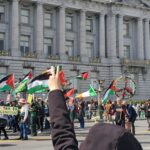 Group of people in front of San Francisco City Hall waving Palestinian flags, with one person holding up a peace sign.