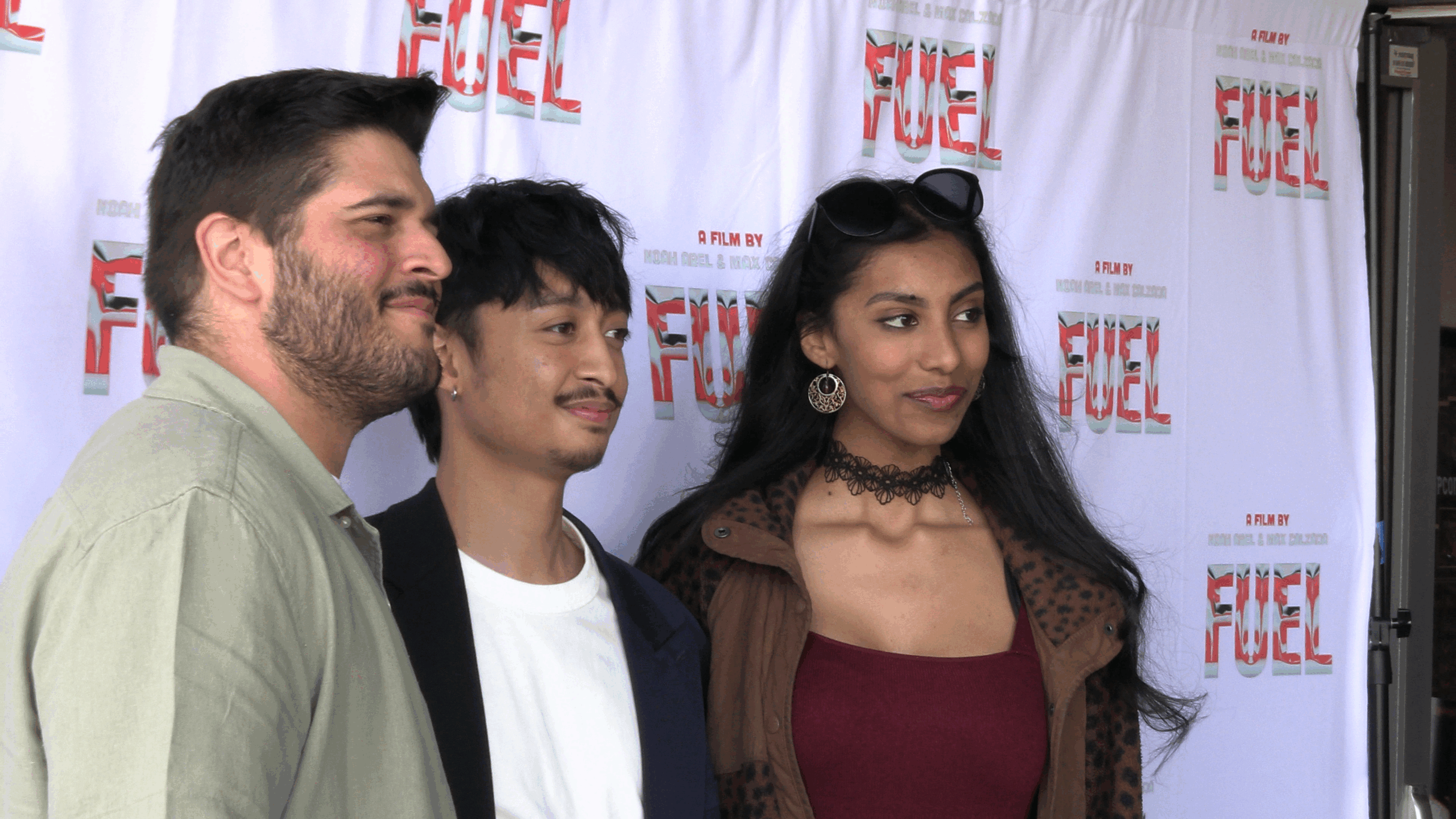 A group of three individuals pose in front of a promotional step-and-repeat banner featuring the stylized text “FUEL” and the phrase “A Film by Noah Abel & Max Calzada.” On the left stands a man with short dark hair and a trimmed beard, wearing a light green button-up shirt. In the center is a young man with short black hair, facial hair, and a small hoop earring, dressed in a white t-shirt and dark blazer. To the right is a woman with long dark hair, wearing sunglasses on her head, large round earrings, a black lace choker, a burgundy top, and a brown coat with a subtle animal print. All three are smiling as they look slightly off-camera, suggesting a posed photo at a film premiere or media event for FUEL.