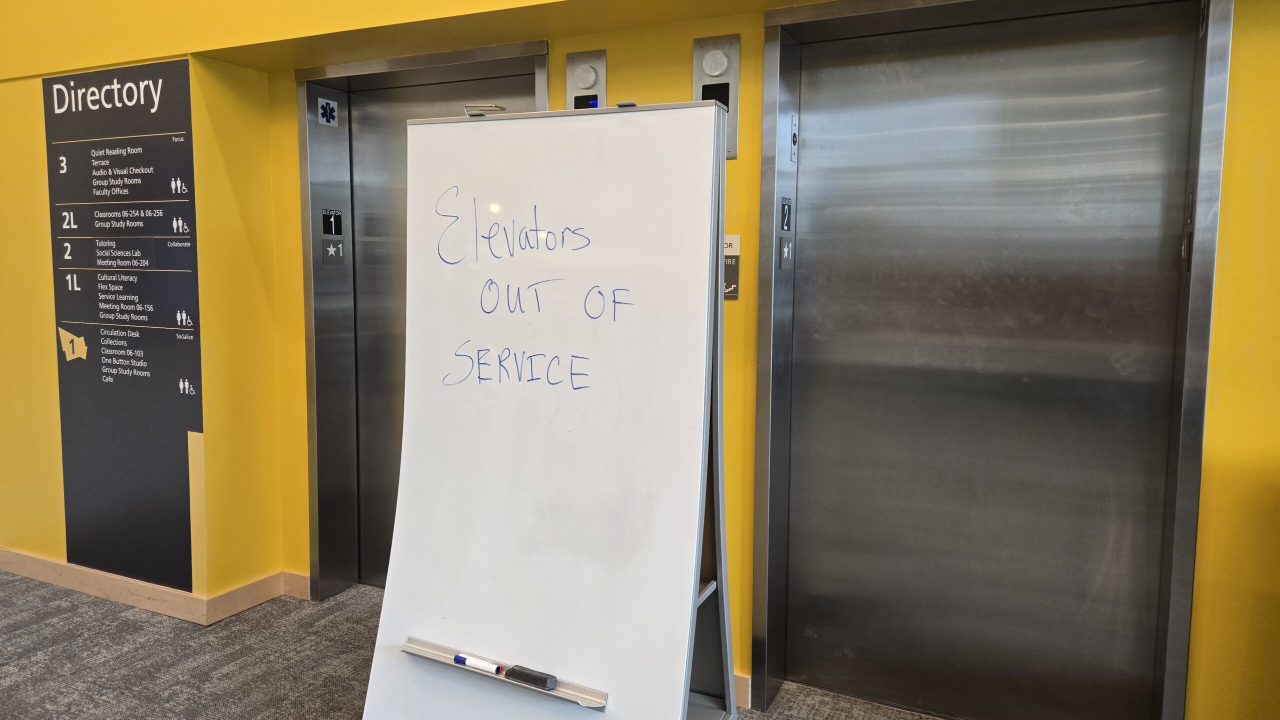A whiteboard with the handwritten message “Elevators out of service” stands in front of two closed elevator doors. The wall behind them is painted yellow, and to the left is a black building directory sign listing floors and rooms, including classrooms, study rooms, and a café. The setting appears to be inside a public or campus building.