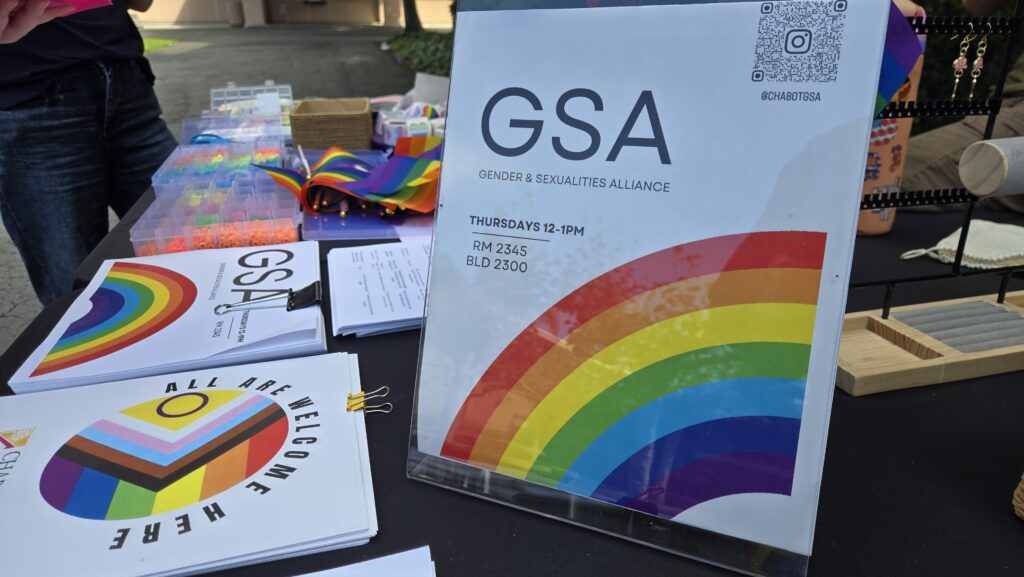 A table display for the Gender & Sexualities Alliance (GSA) featuring a sign with a rainbow design that reads “GSA – Gender & Sexualities Alliance, Thursdays 12–1 PM, RM 2345, BLD 2300.” The table includes informational pamphlets, rainbow-colored materials, and craft supplies. A QR code and the Instagram handle @chabotgsa are visible on the sign. The setup appears to be part of an outdoor campus event.