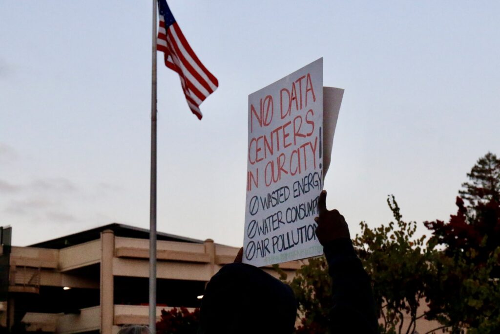 A person raises a “No data centers in our city!” sign beneath an American flag.