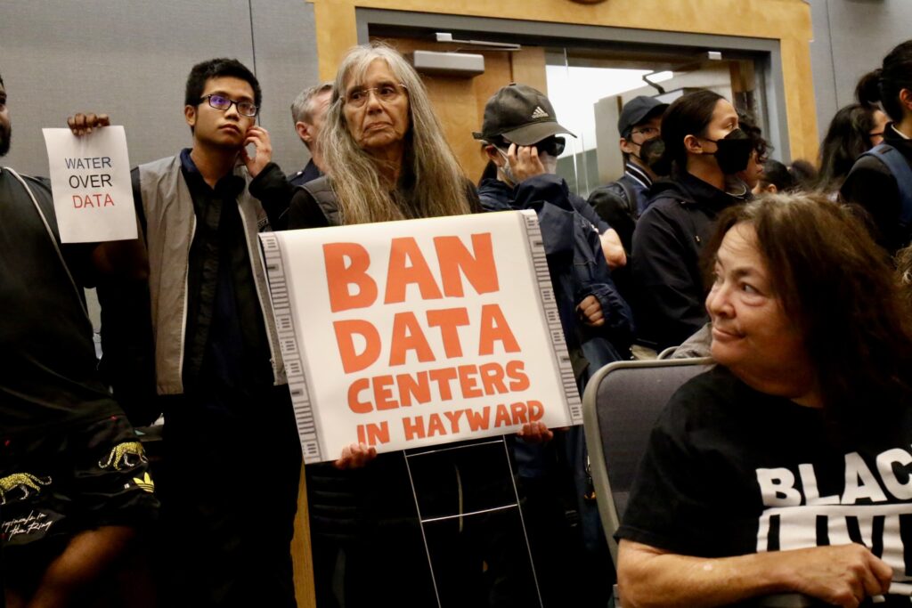 People in a crowded room hold signs reading “Ban data centers in Hayward” and “Water over data.”